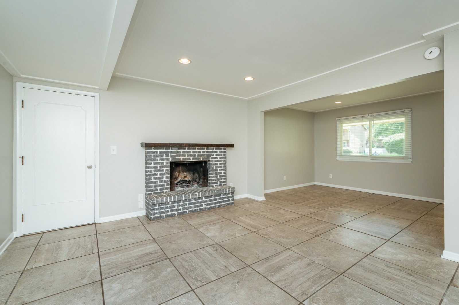 Empty living room with a brick fireplace, large tile floors, and an adjoining windowed room.
