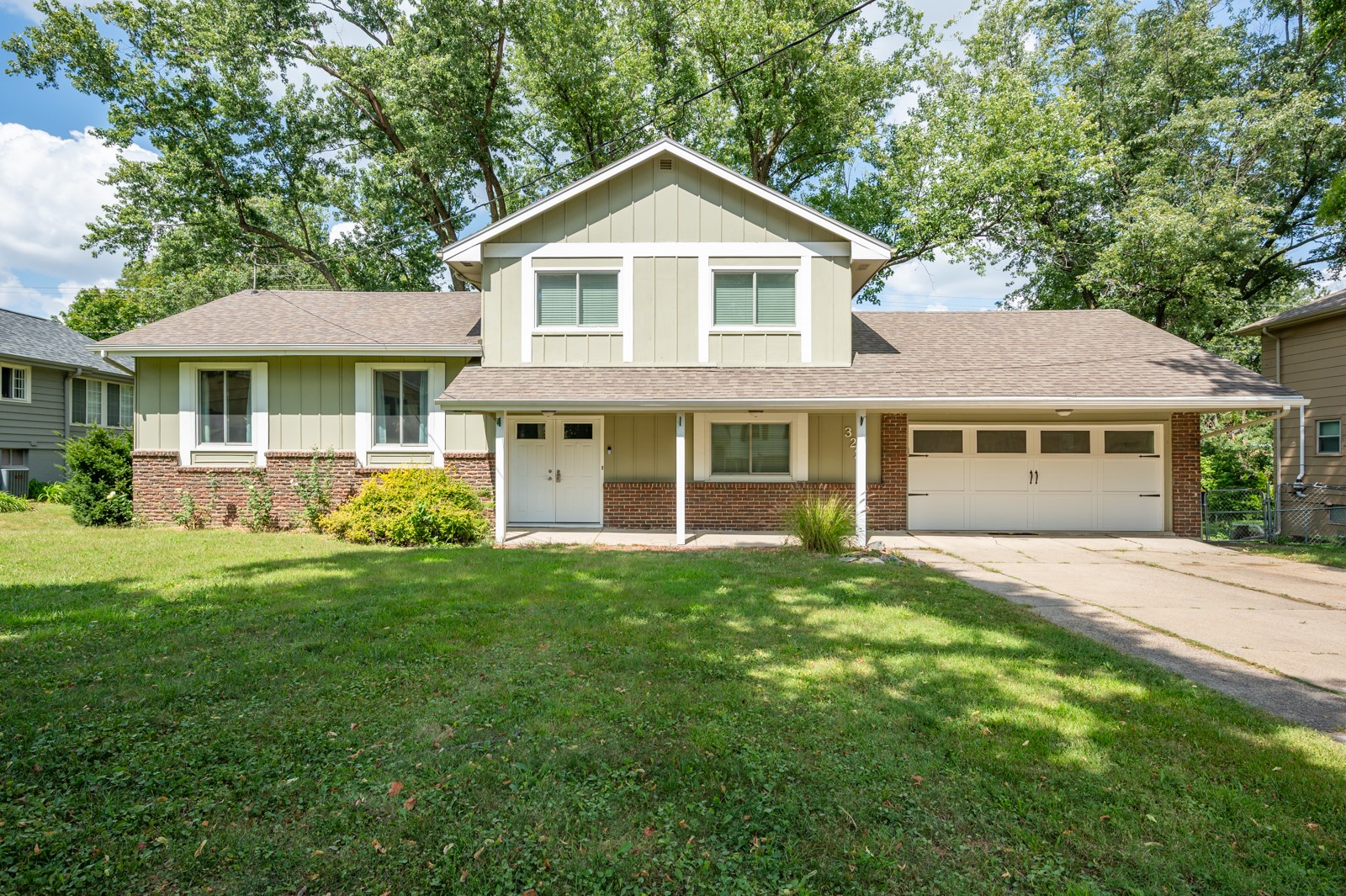 Two-story pale green house with brick lower facade, attached garage, and shaded front lawn.