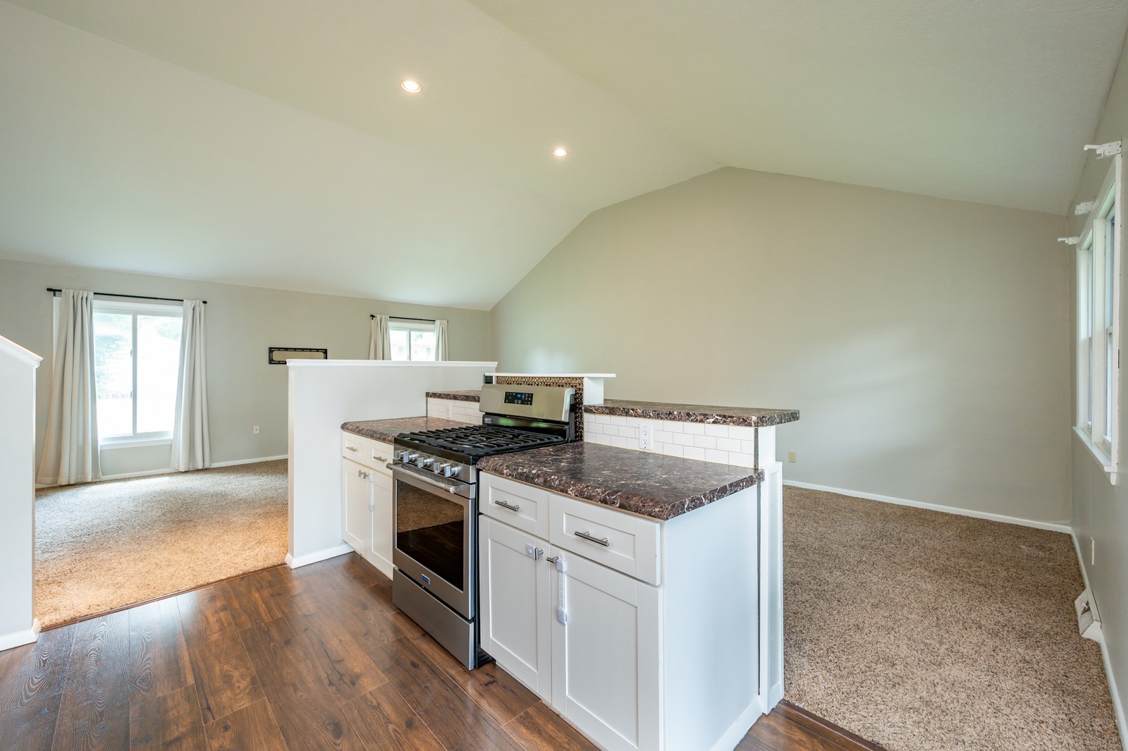 Vacant kitchen with white cabinets, dark countertops, stainless steel gas range, and adjoining carpeted living area.