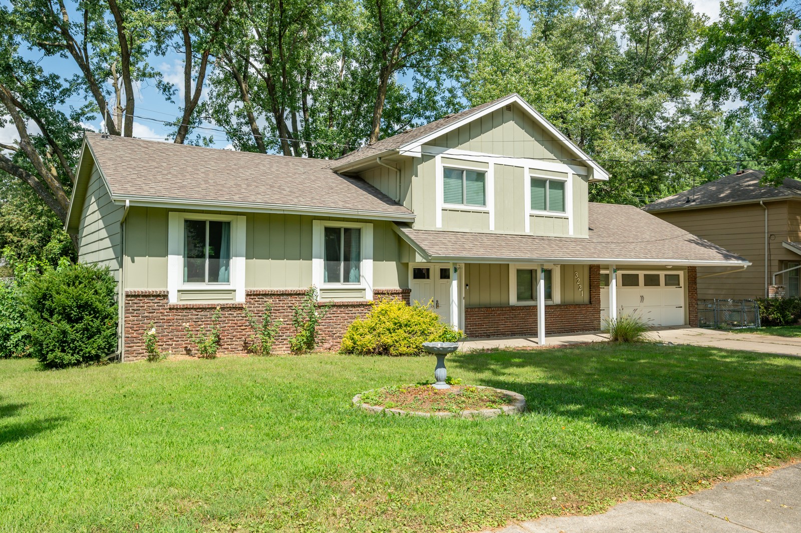 Two-story pale green house with brick lower walls, attached garage, mature trees, and a broad front lawn.