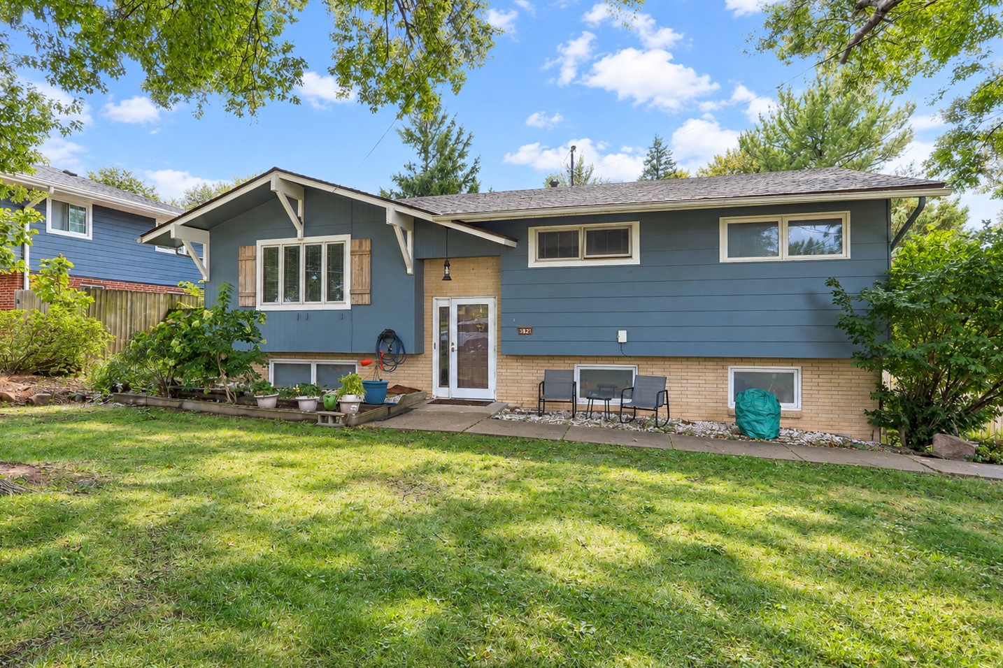 Blue split-level house with brick lower facade, patio seating, and a leafy garden.