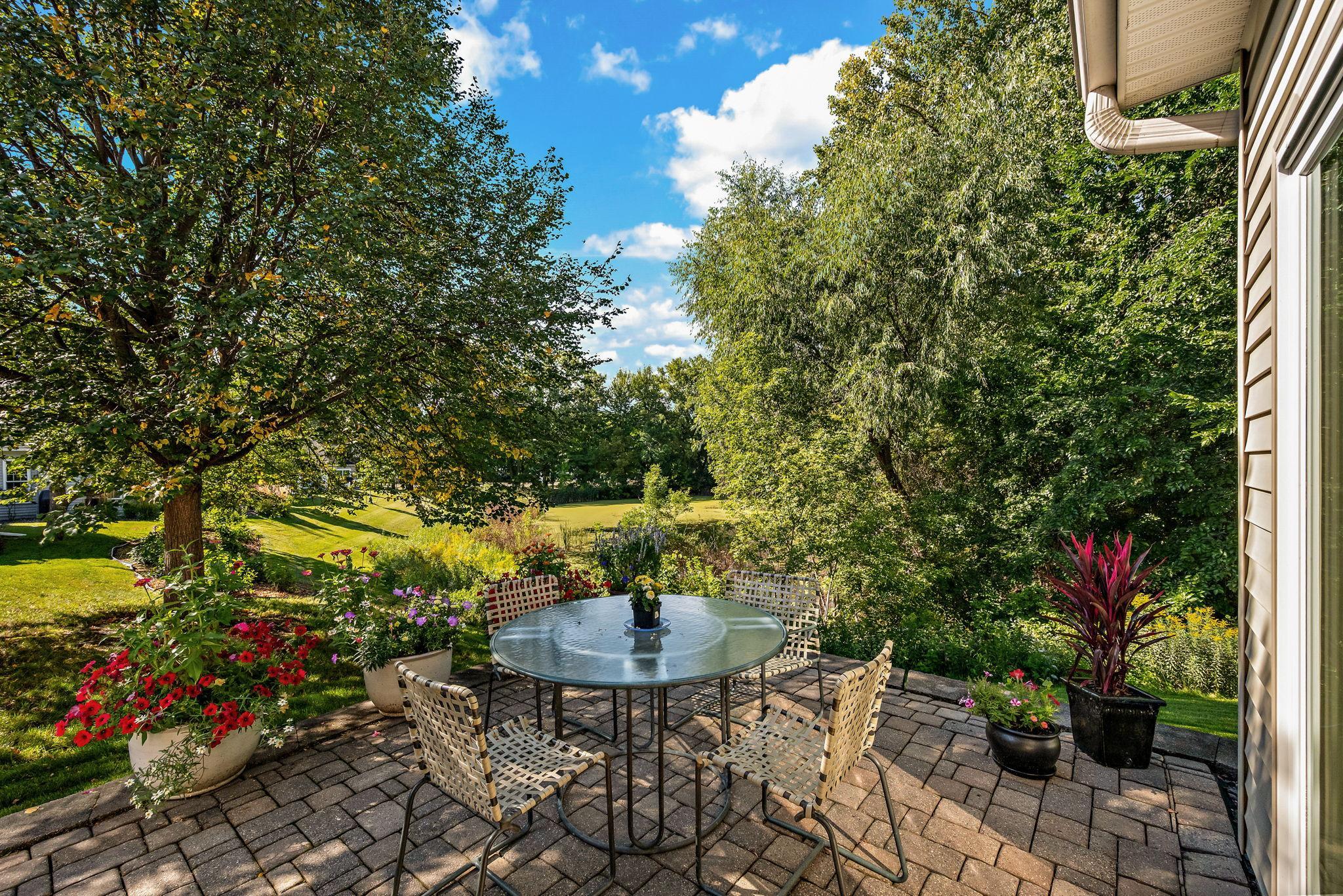 Brick patio with a glass dining table and woven chairs amid colorful flowers and mature trees.