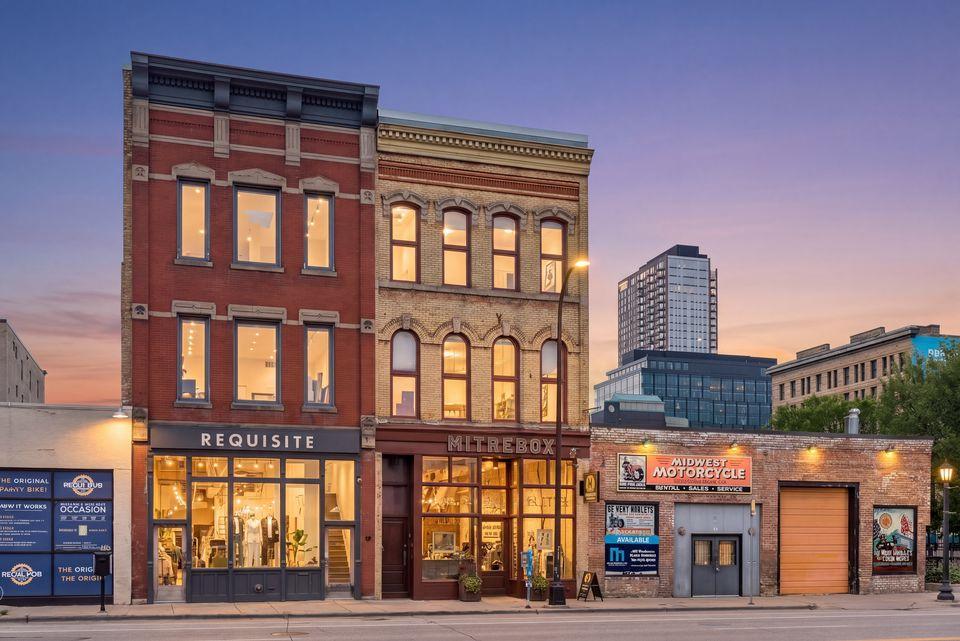 Historic brick storefronts at dusk, with illuminated shops and modern city buildings behind them.