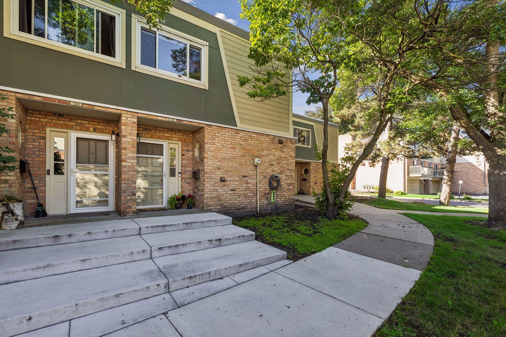 Attached brick-and-siding townhouse entrances beside a curved sidewalk and mature trees.