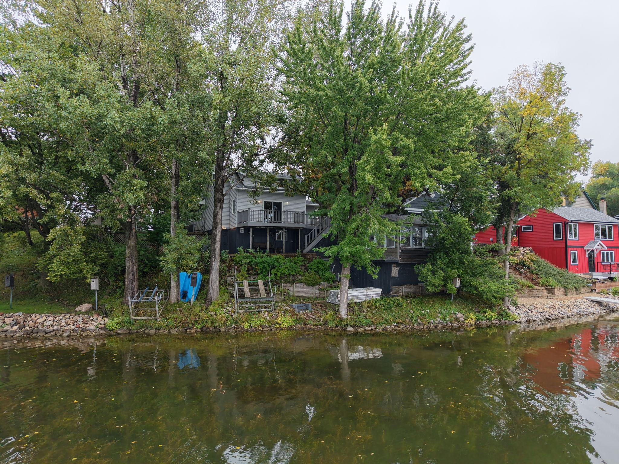 Gray lakeside house with a raised deck, framed by trees beside a rocky shore.