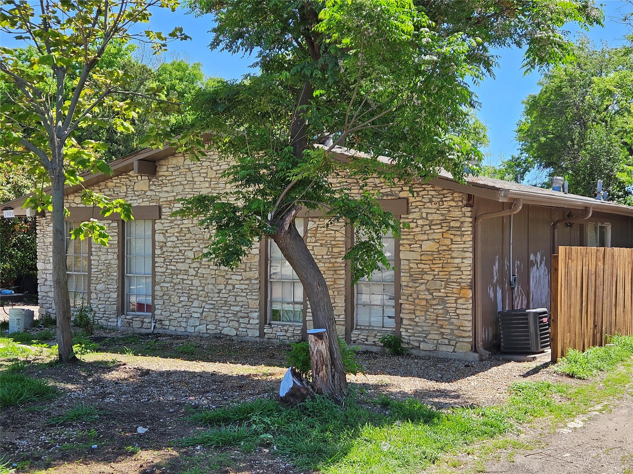 Stone ranch house shaded by mature trees, with gravel landscaping and a side air conditioner.