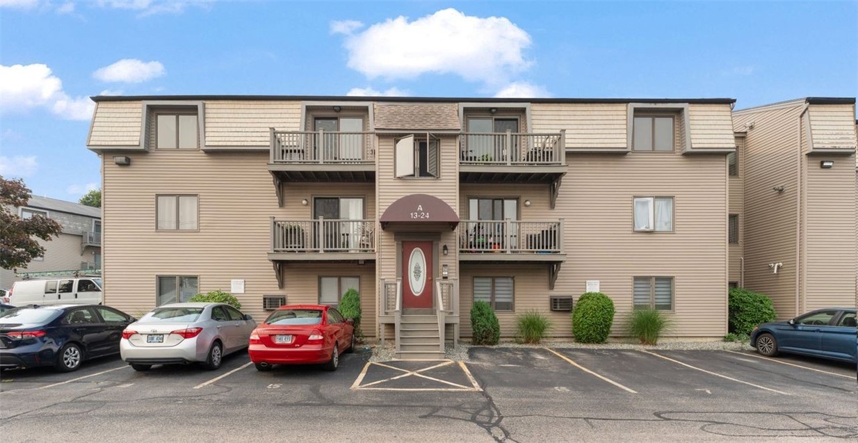 Beige townhouse building with several balconies, a red entrance door, and cars parked in front.