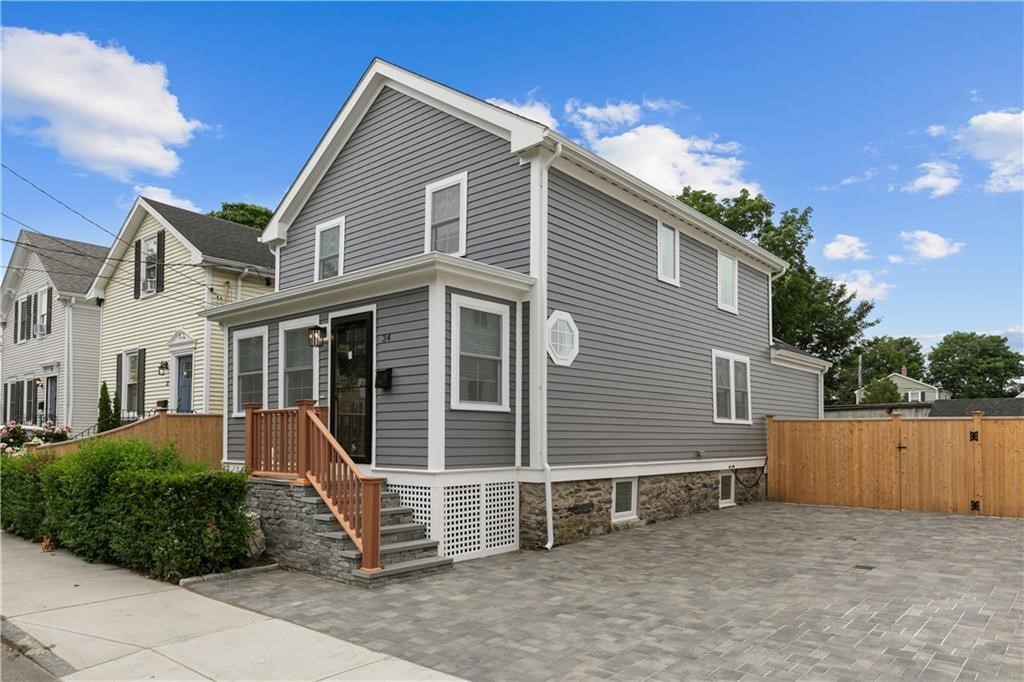 Gray two-story house with a stone foundation, front porch, and fenced paved courtyard.
