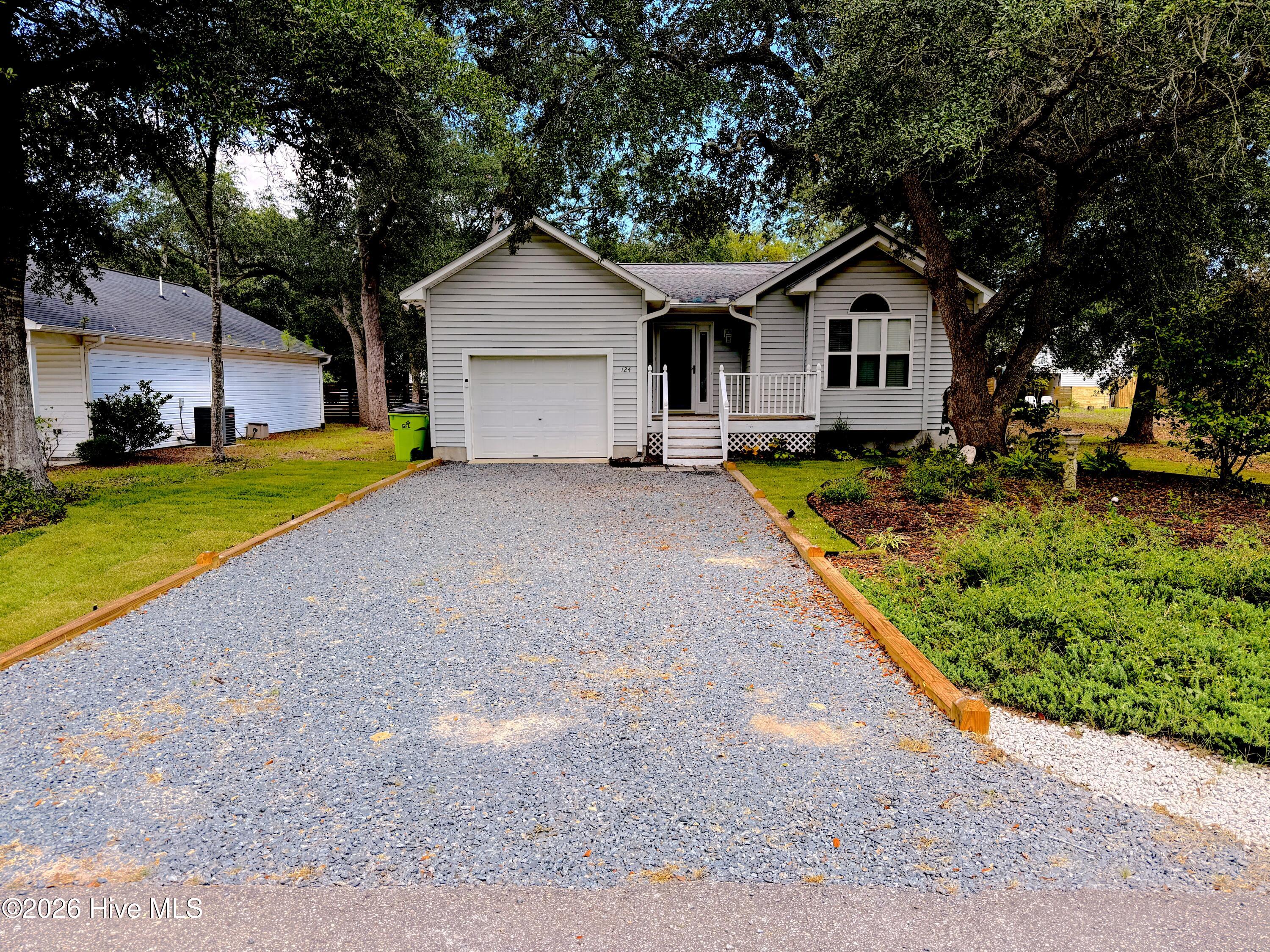 Gray single-story home with a covered porch, garage, and wide gravel driveway beneath mature trees.