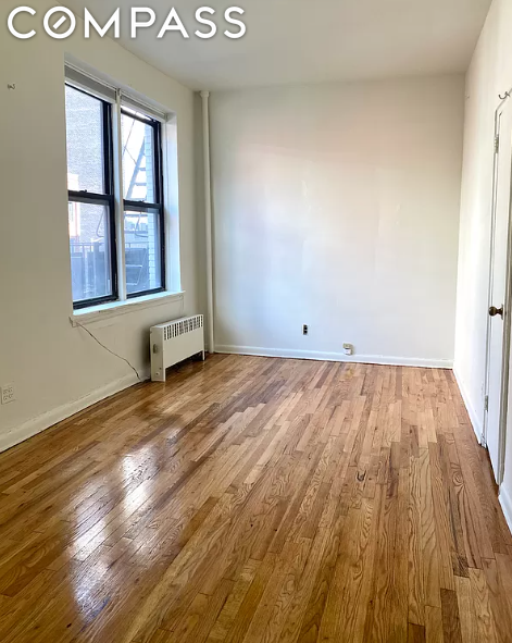 Vacant apartment room with polished hardwood floors, a large window, and a radiator.