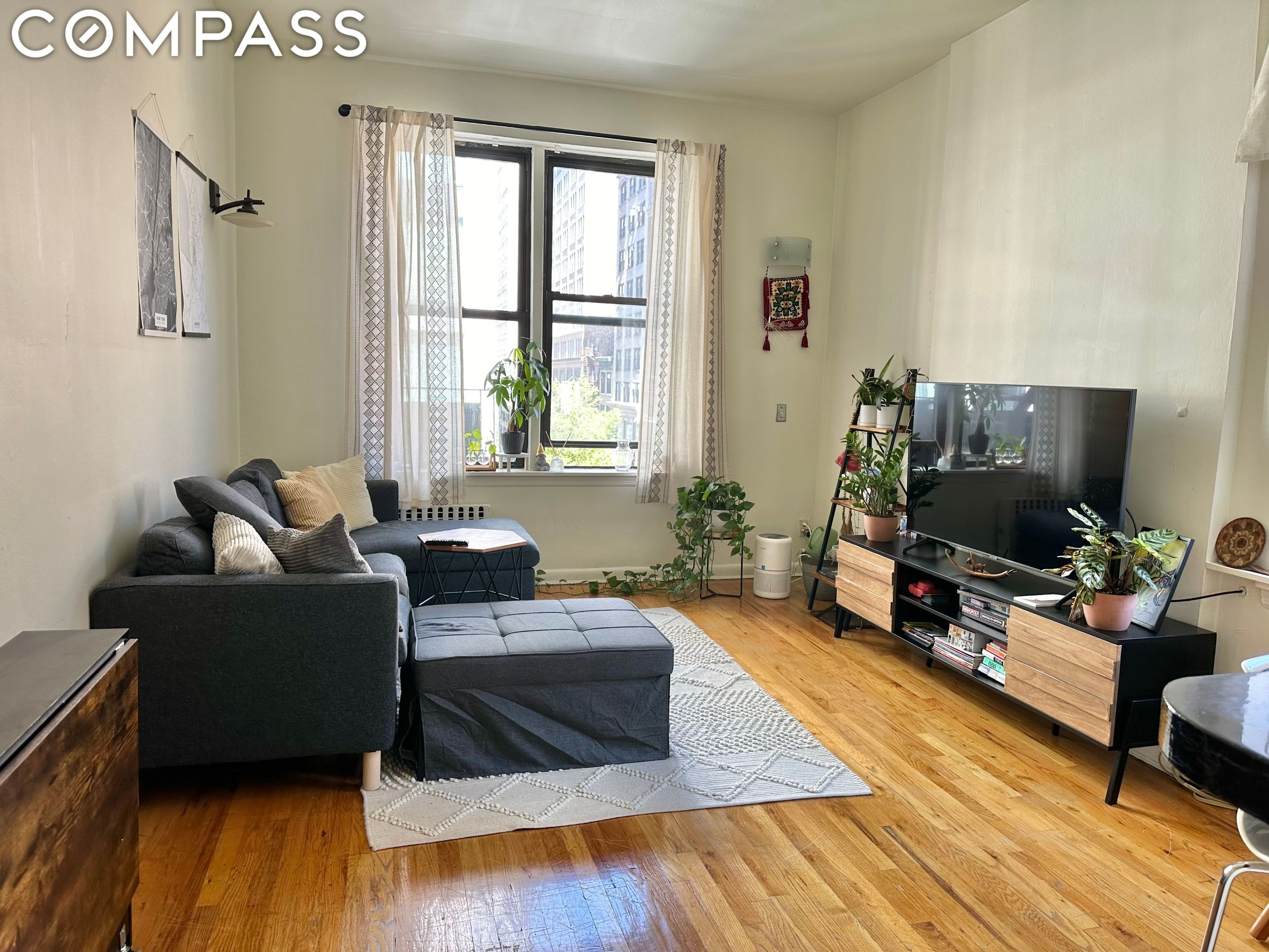 Furnished living room with gray sectional, large windows, hardwood floors, and city buildings outside.
