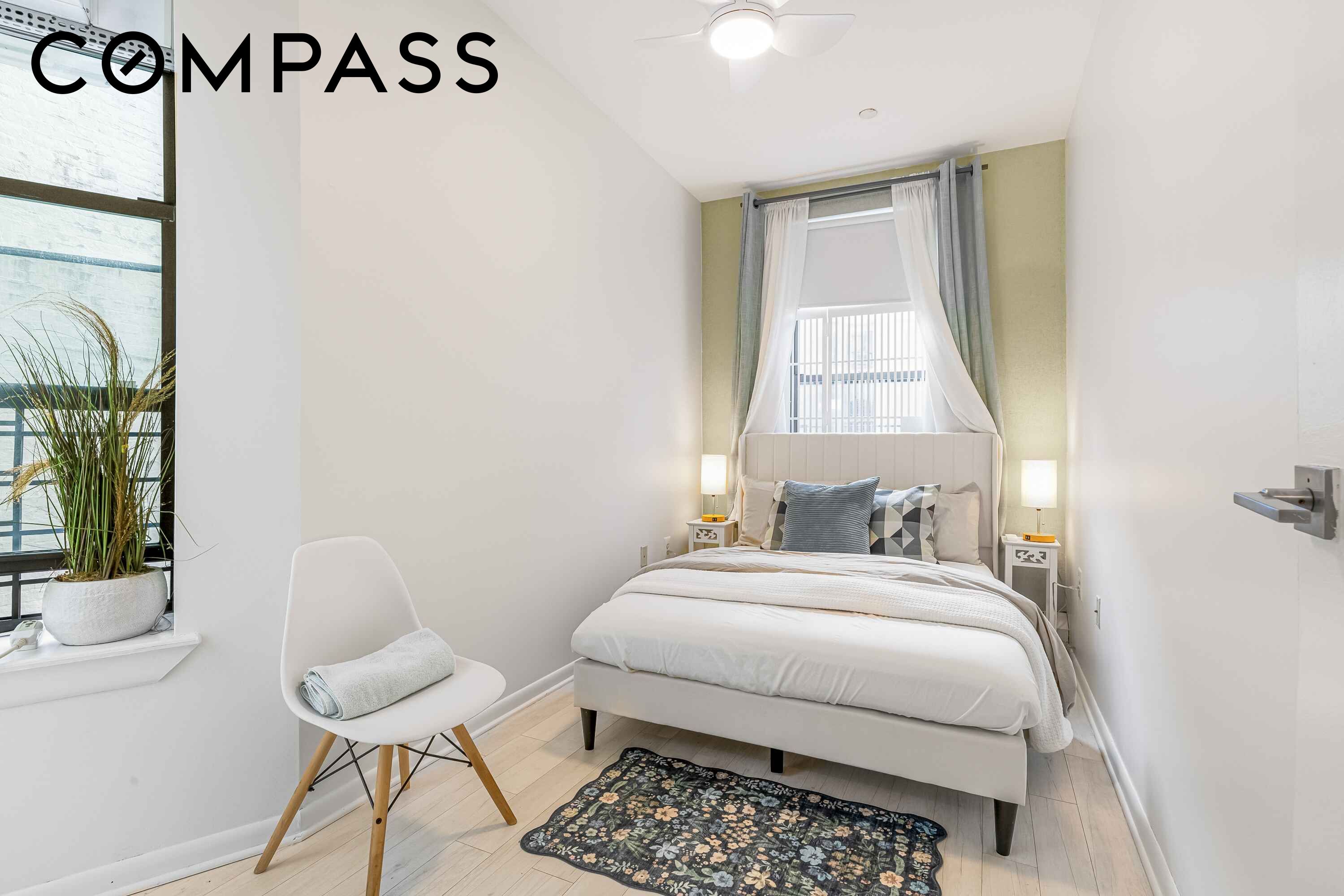 Staged bedroom with a white bed beneath a curtained window and a chair beside a tall window.