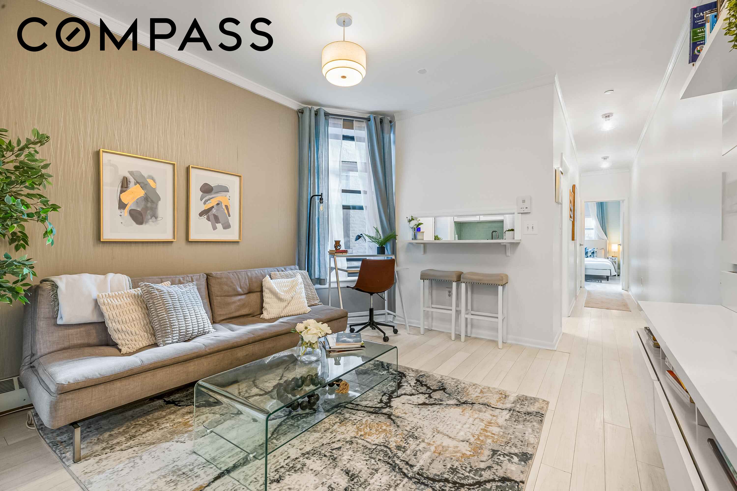 Staged living room with a gray sofa, glass coffee table, and breakfast bar opening to a hallway.