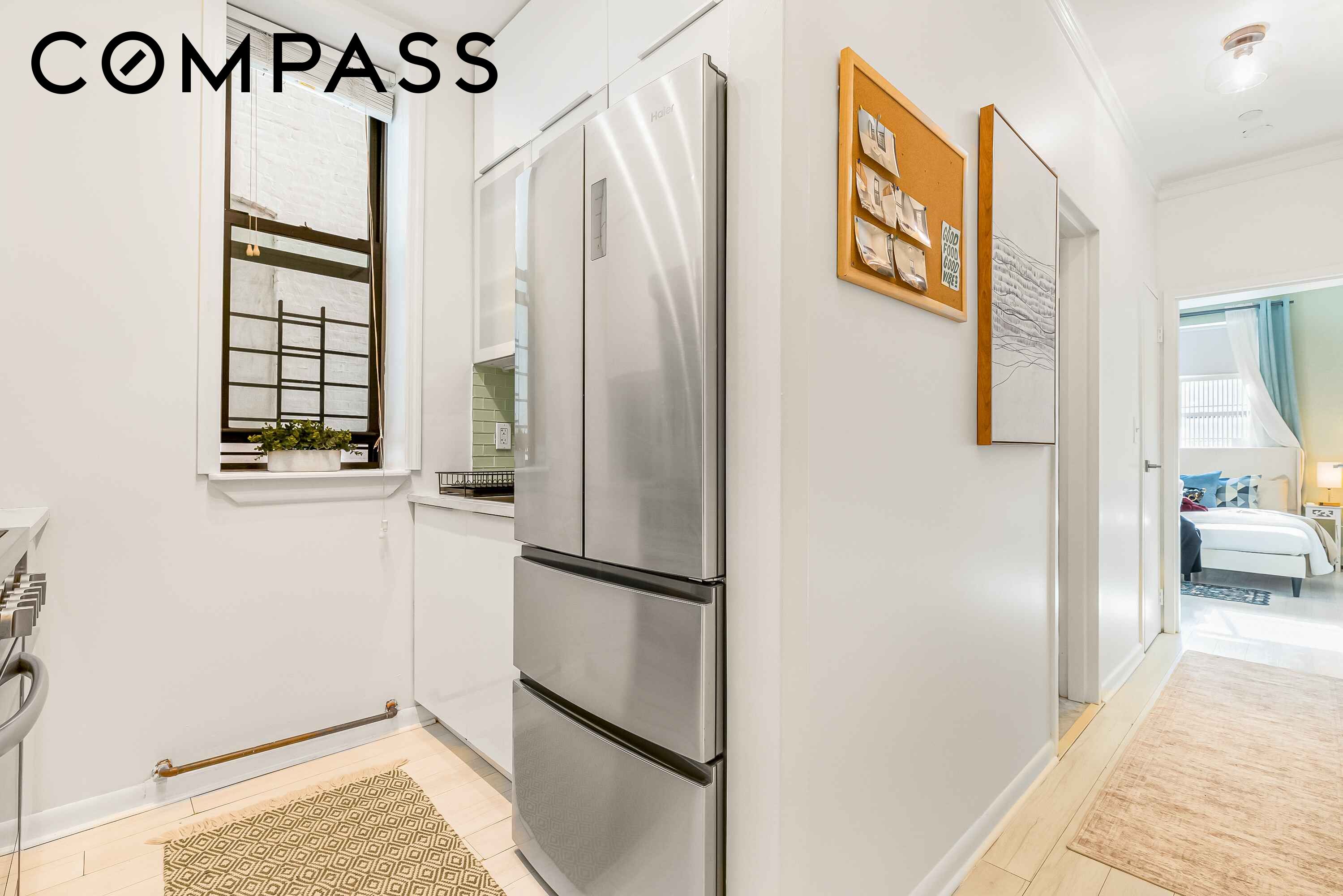 Bright apartment kitchen with a stainless steel refrigerator and a bedroom visible through the hallway.
