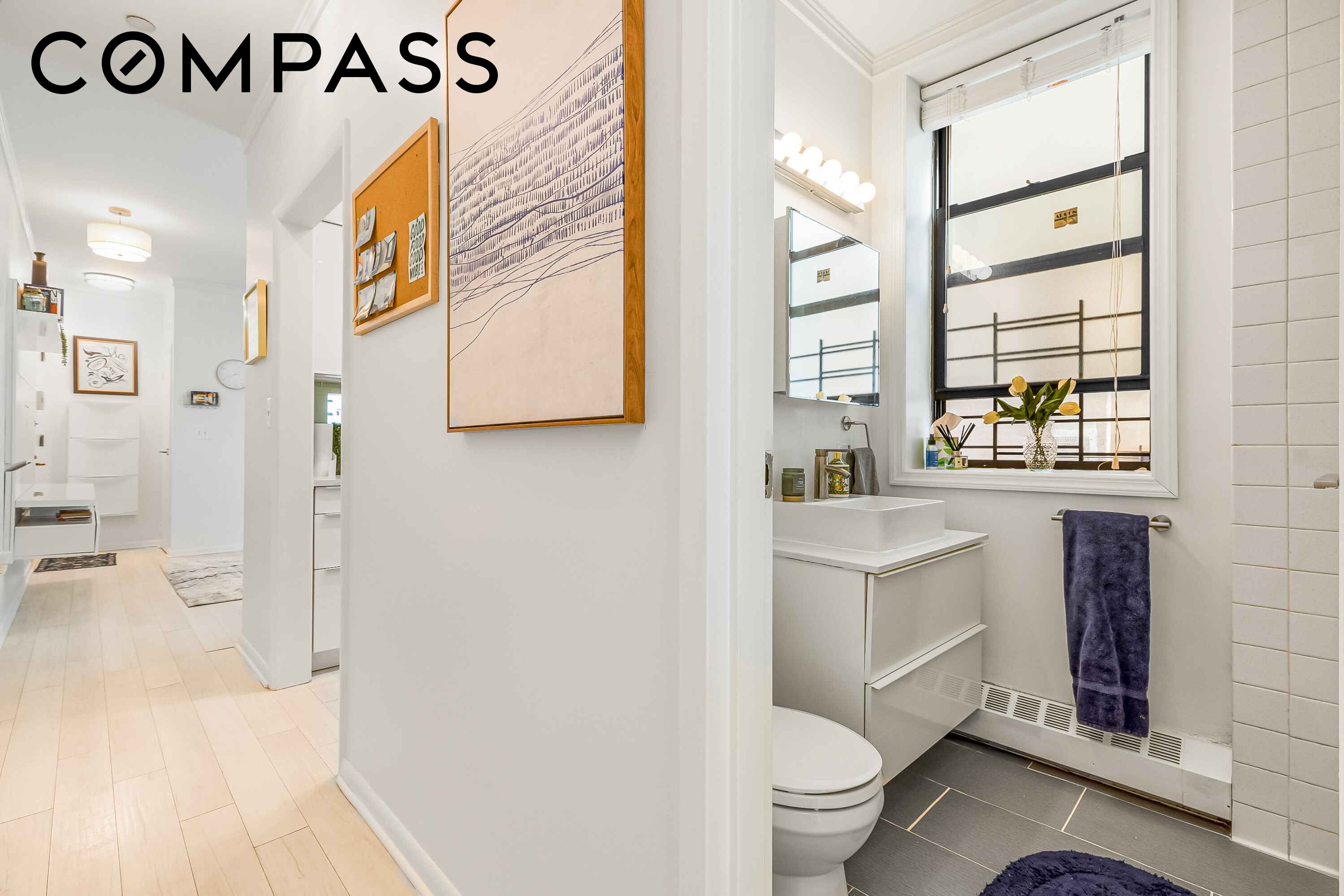 Bathroom with a vessel sink beneath a large window, dark tiled floor, and a purple towel beside an art-lined hallway.