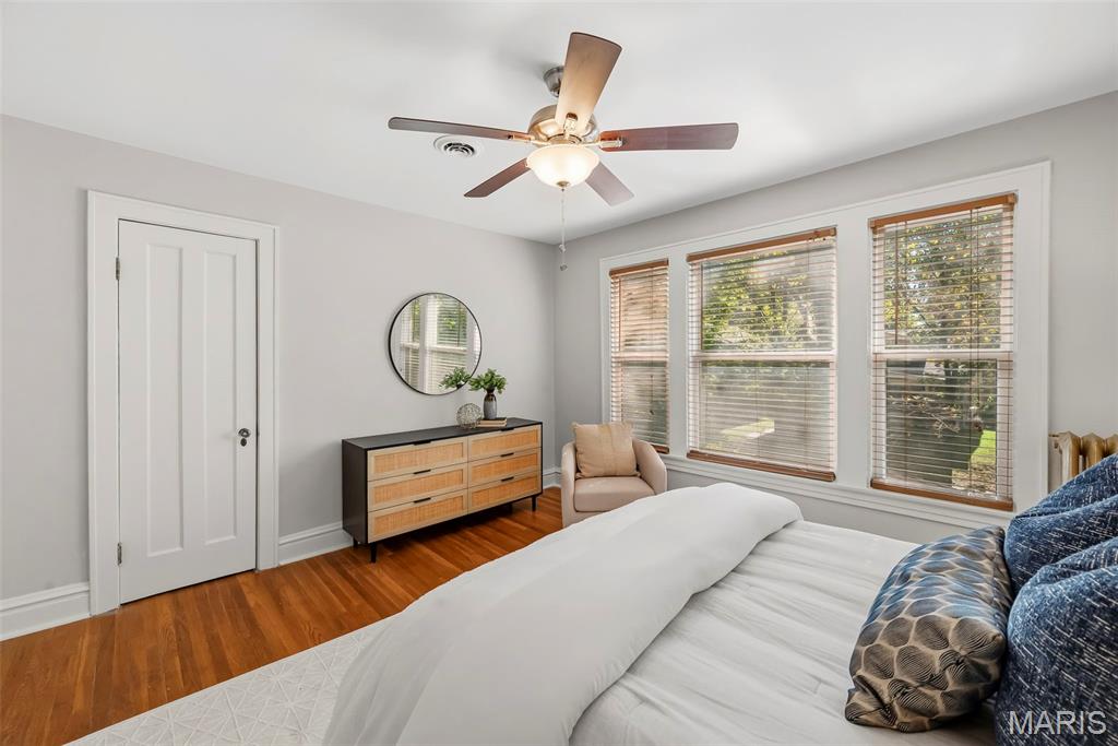 Bedroom with hardwood floors, a ceiling fan, and three large windows overlooking a green yard.