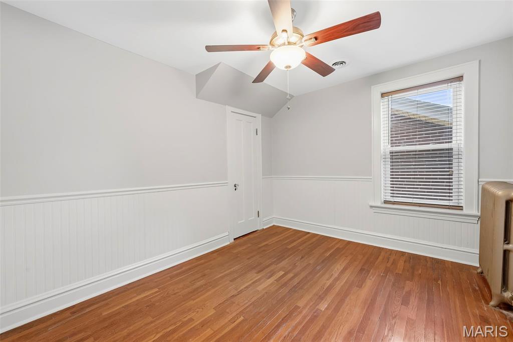 Empty bedroom with hardwood floors, white wainscoting, a ceiling fan, and a window with blinds.