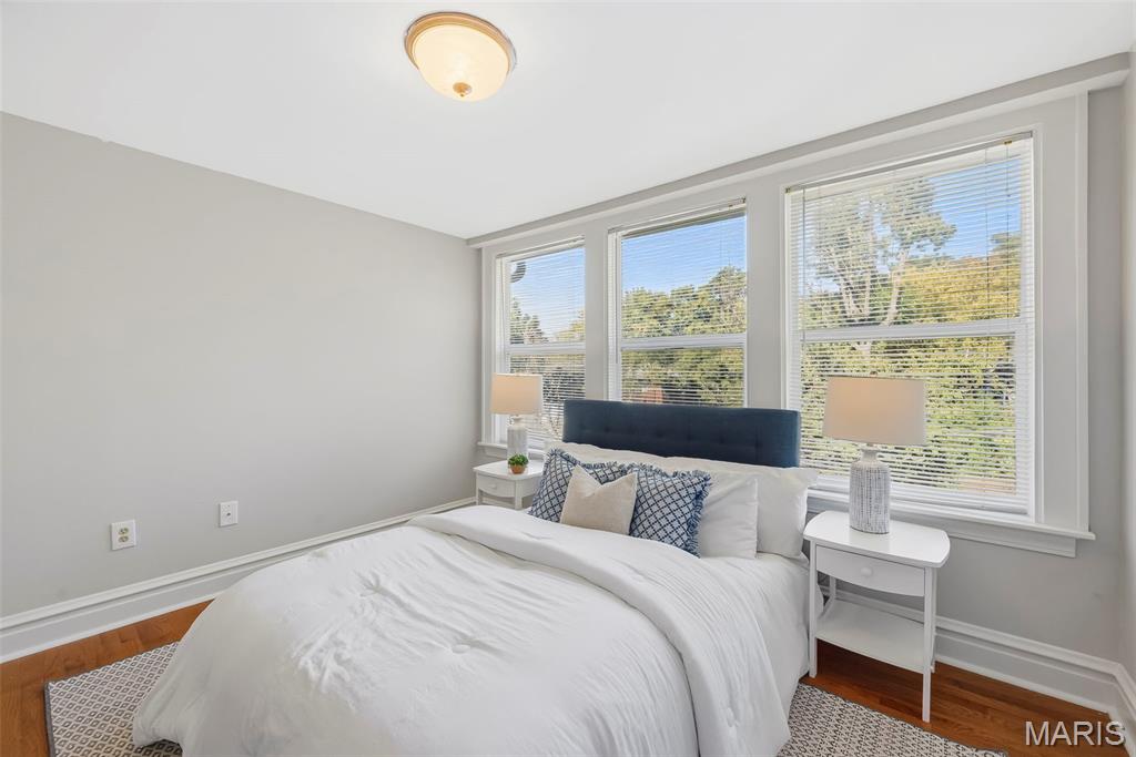 Staged bedroom with a white bed, navy headboard, and three large windows overlooking trees.
