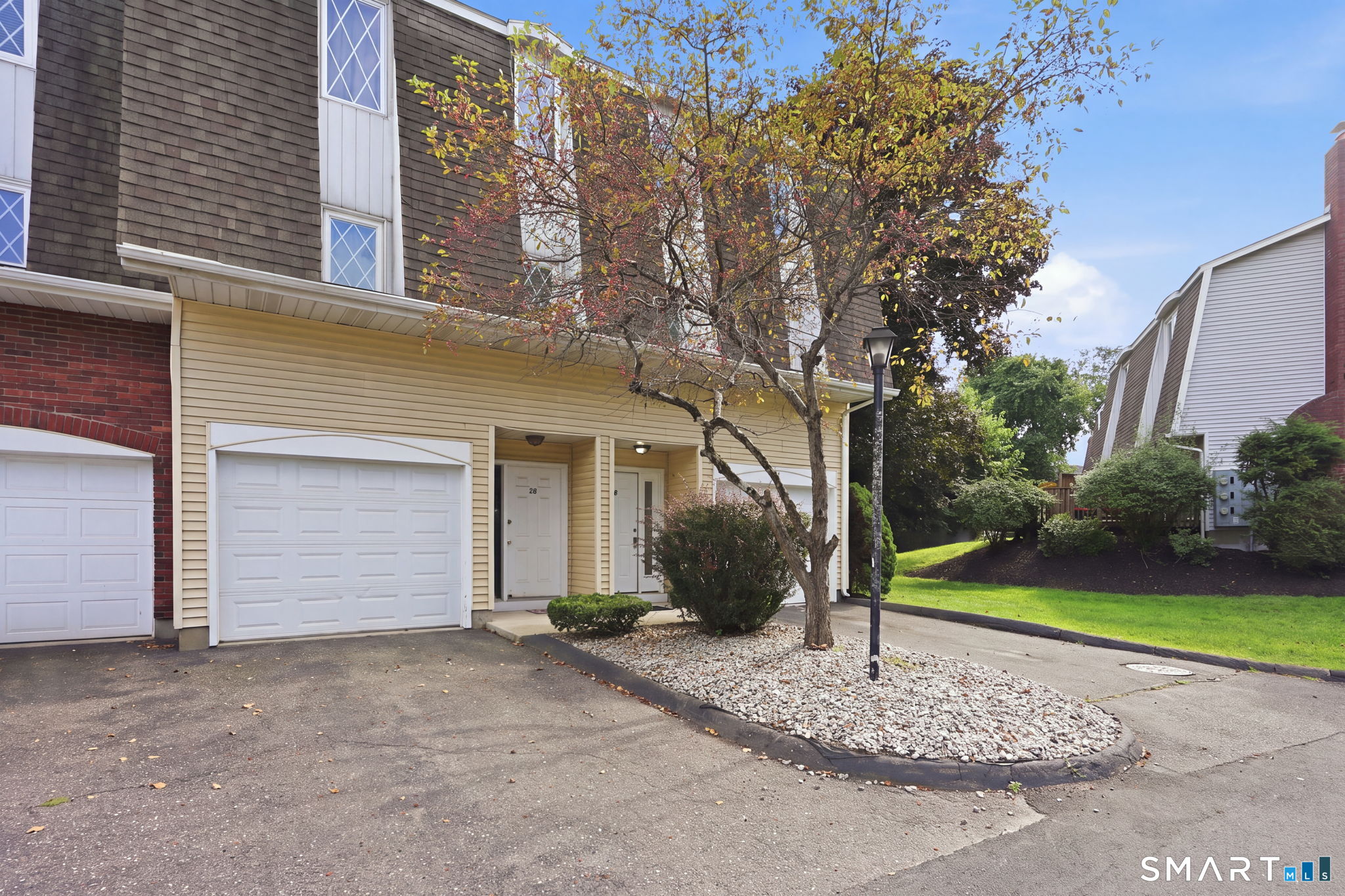 Attached townhouse with a white garage door, landscaped stone bed, and autumn tree.