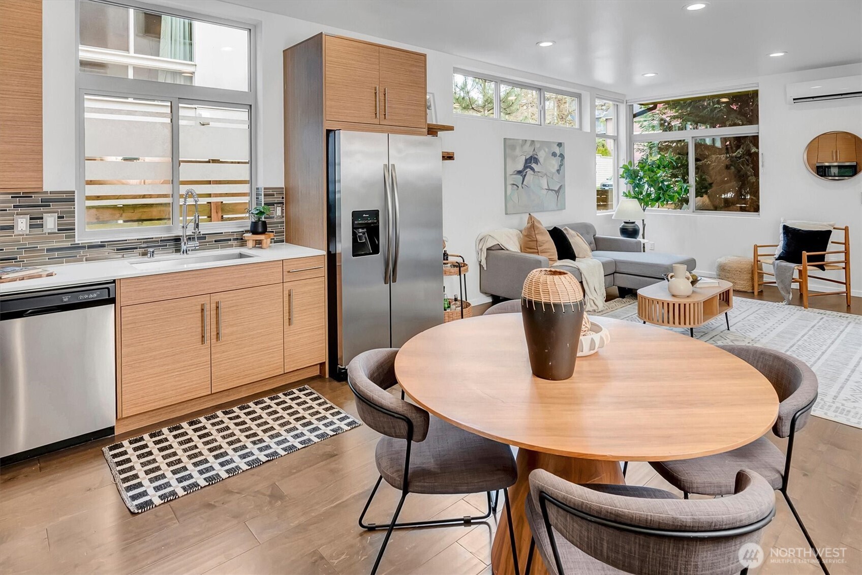 Open-plan kitchen and living room with light wood cabinets, a round dining table, and large windows.