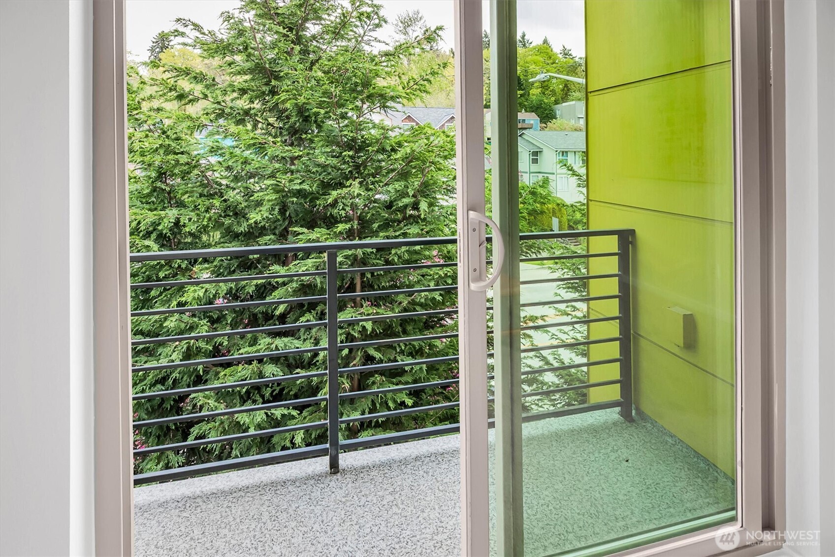 Apartment balcony with a black railing, bright green privacy wall, and leafy trees beyond.