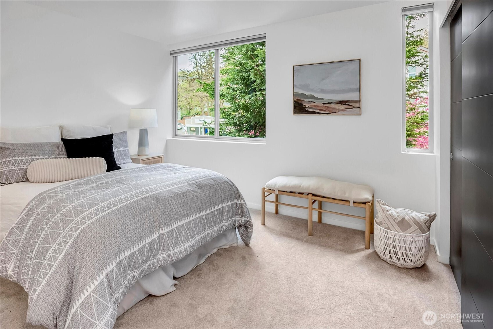 Furnished bedroom with a patterned gray bedspread, bench, and windows overlooking leafy trees.