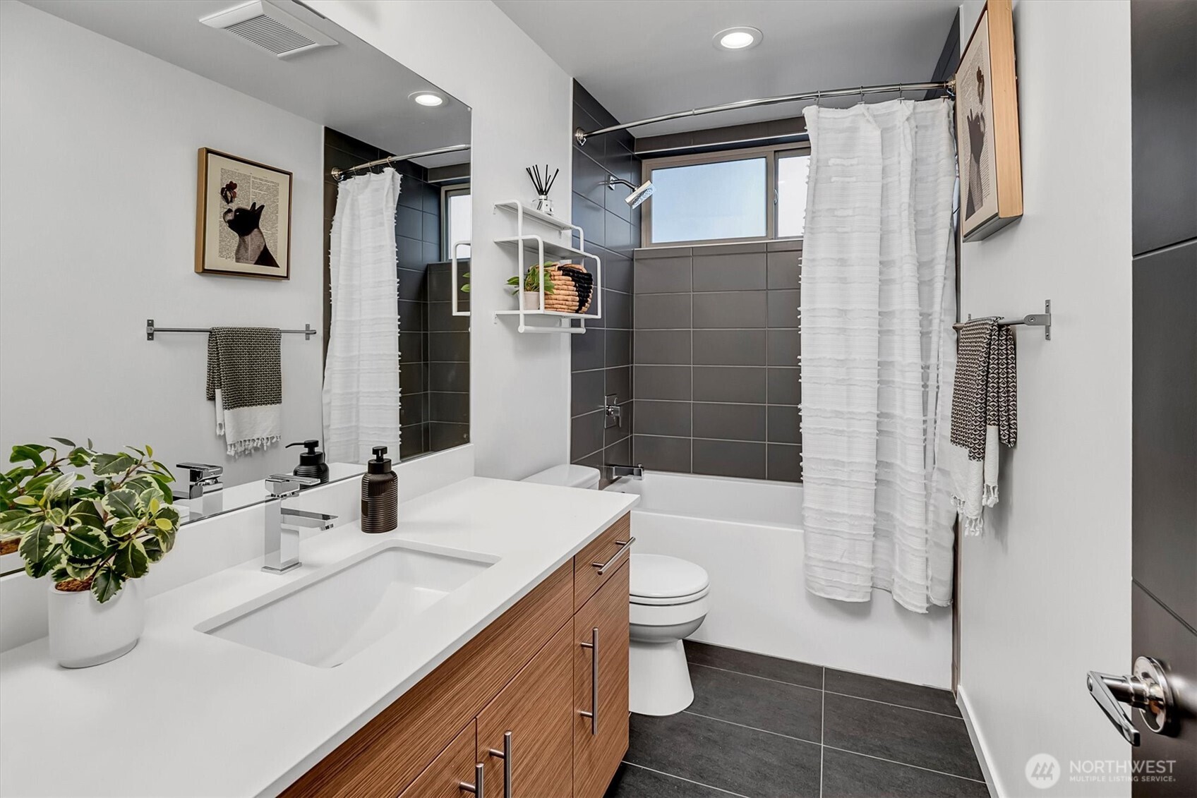 Modern bathroom with a wood-grain vanity, gray tile, and a tub beneath a window.