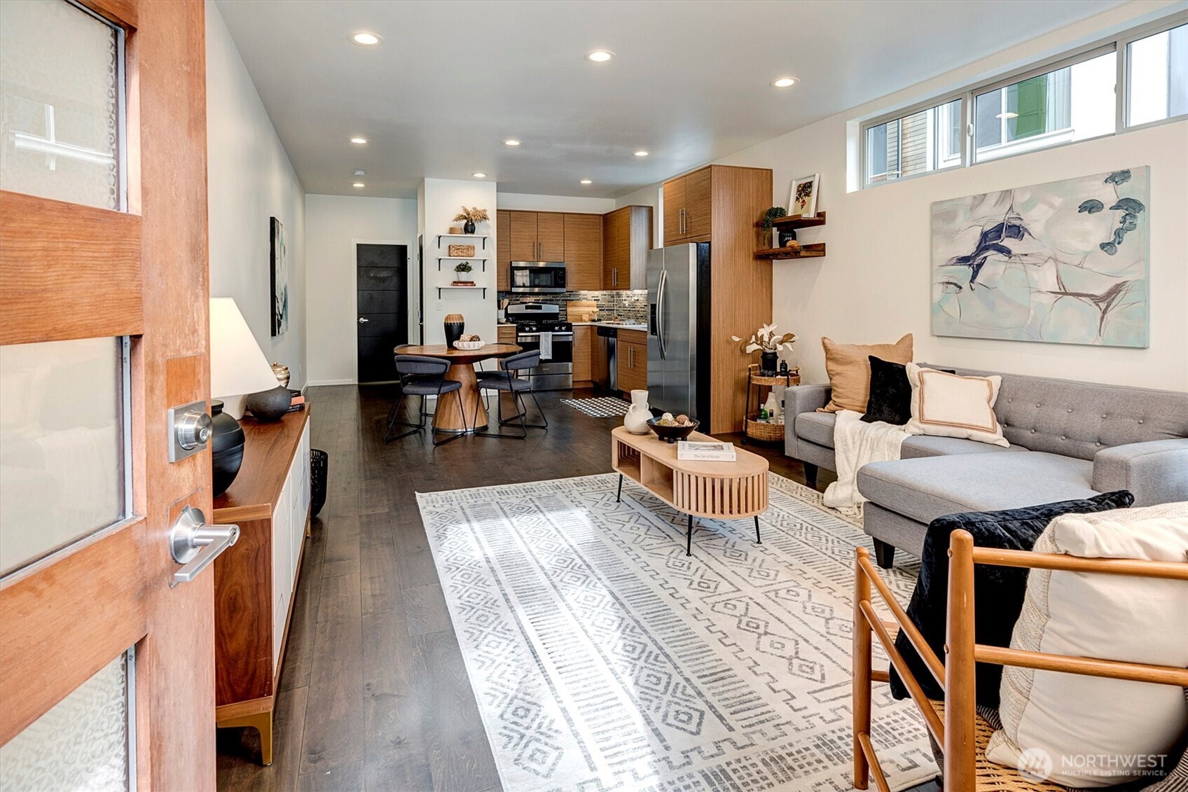 Open-plan living room and kitchen with warm wood cabinets, gray sectional, and patterned rug.
