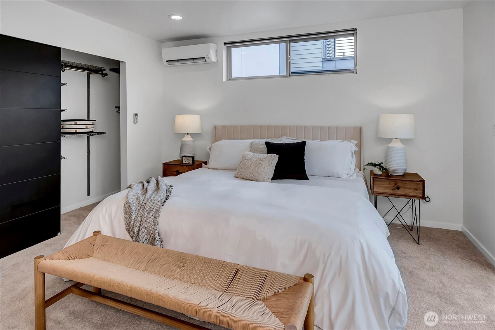 Staged bedroom with a white bed, woven bench, open closet, and high window.