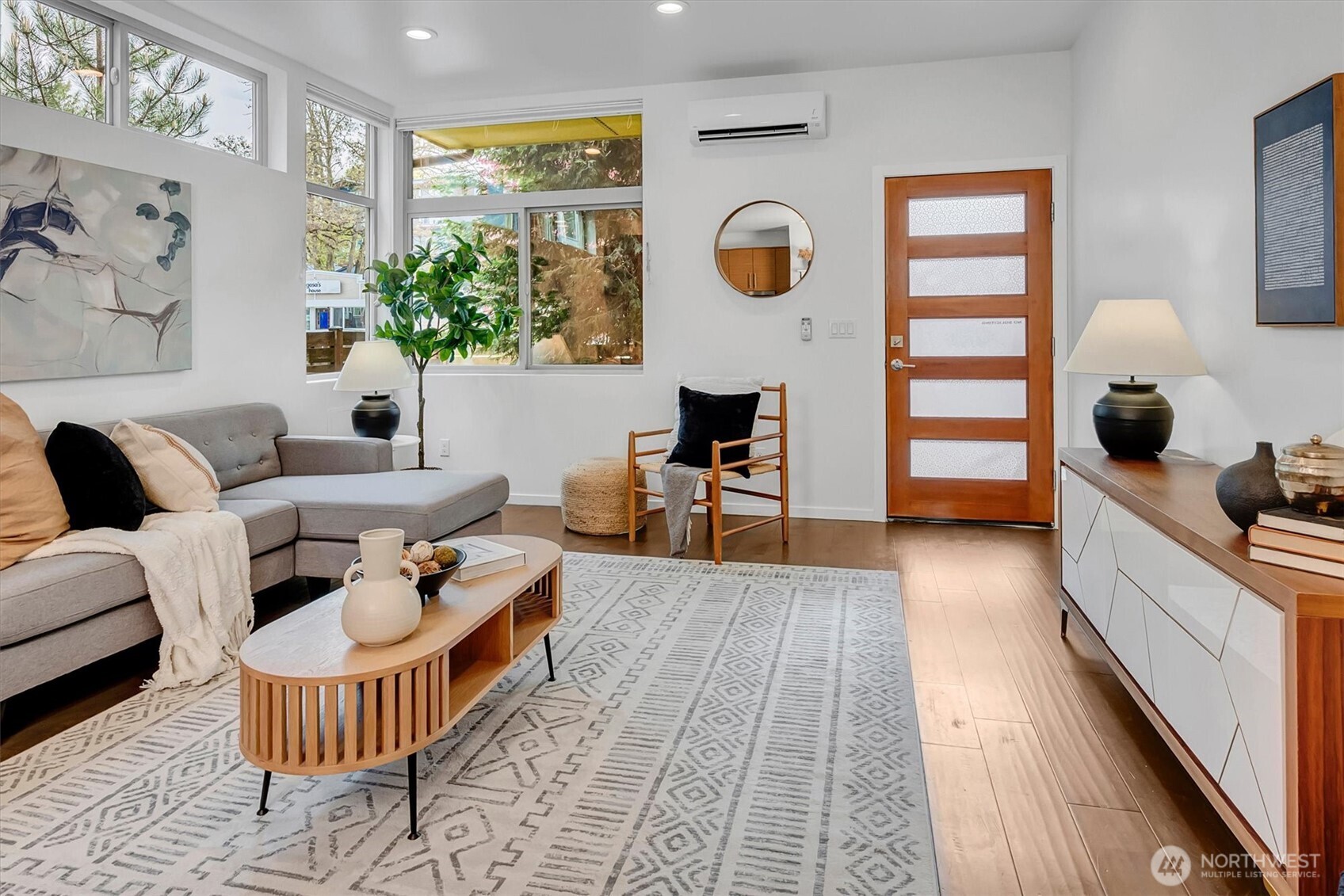 Staged living room with a gray sectional, patterned rug, large windows, and a wood entry door.