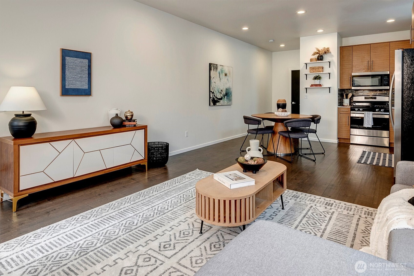 Open-plan living and dining area with a patterned rug, round wood table, and warm wood kitchen cabinets.