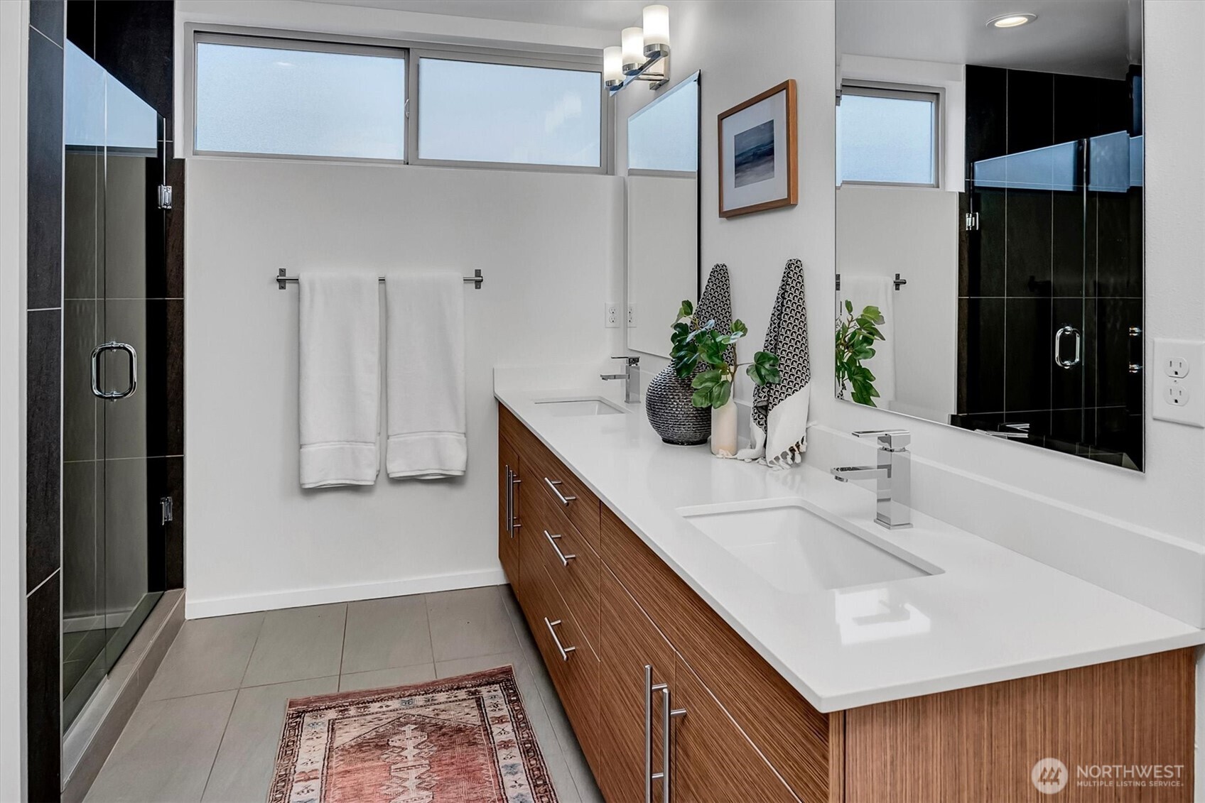 Modern bathroom with a long wood-grain double vanity, white counters, and a glass-enclosed dark-tile shower.