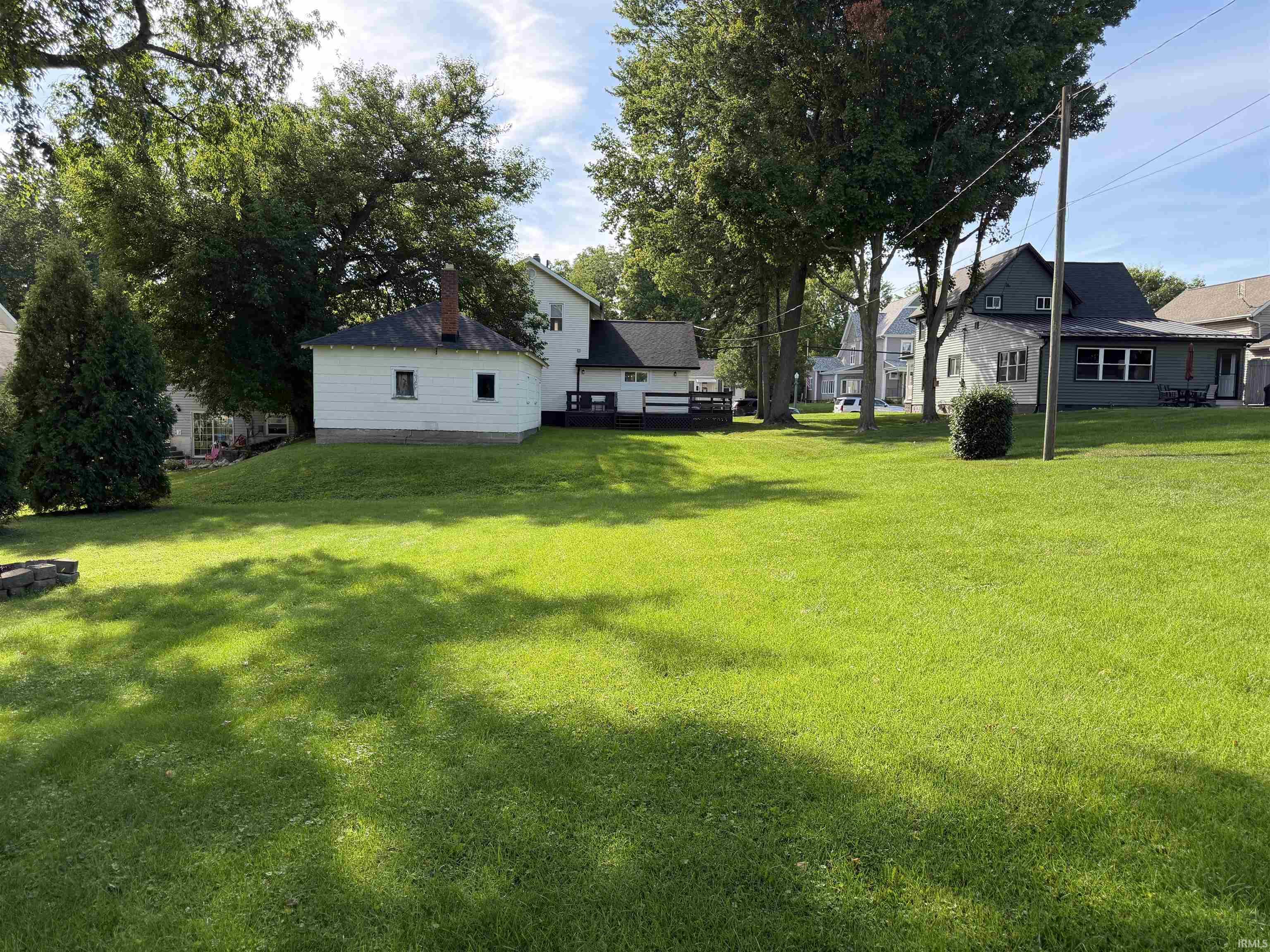 Large grassy backyard with mature trees, a gray-sided house and raised deck, and a small white outbuilding.
