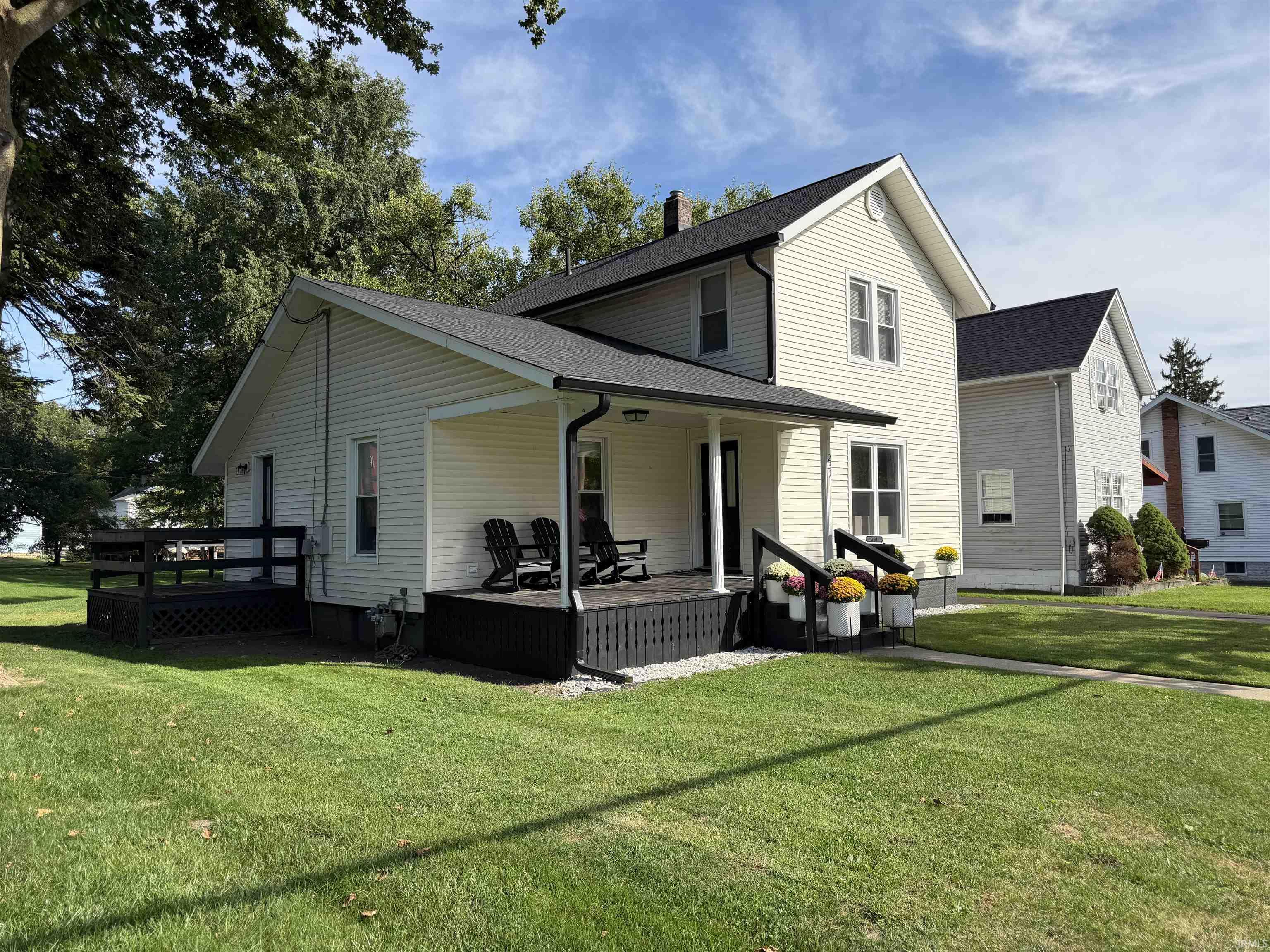 Two-story cream house with a covered porch, black railings, potted flowers, and a broad green lawn.