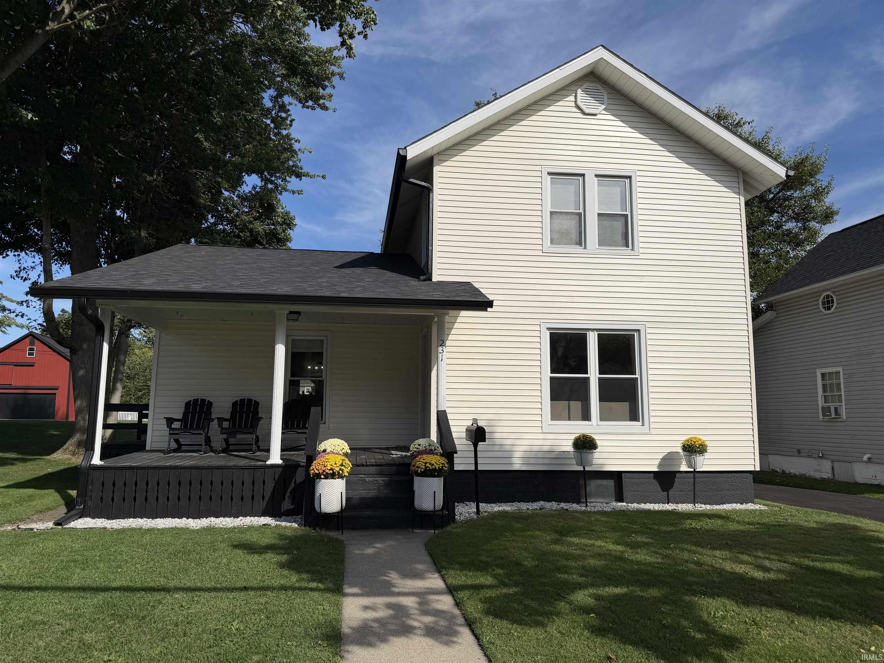 Two-story cream-sided house with a covered porch, Adirondack chairs, and flower planters.