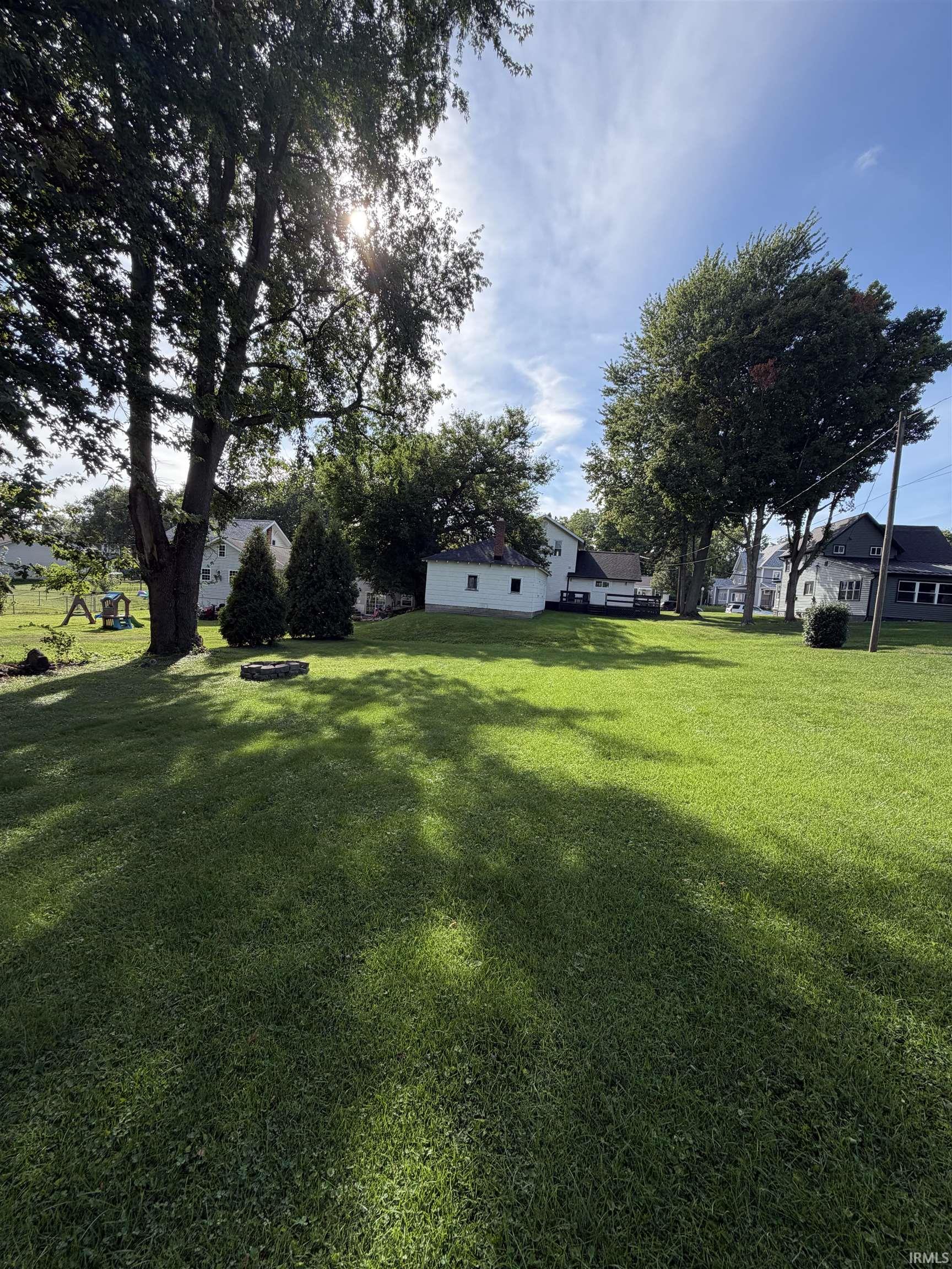Spacious backyard with a green lawn, mature shade trees, and a small fire pit.