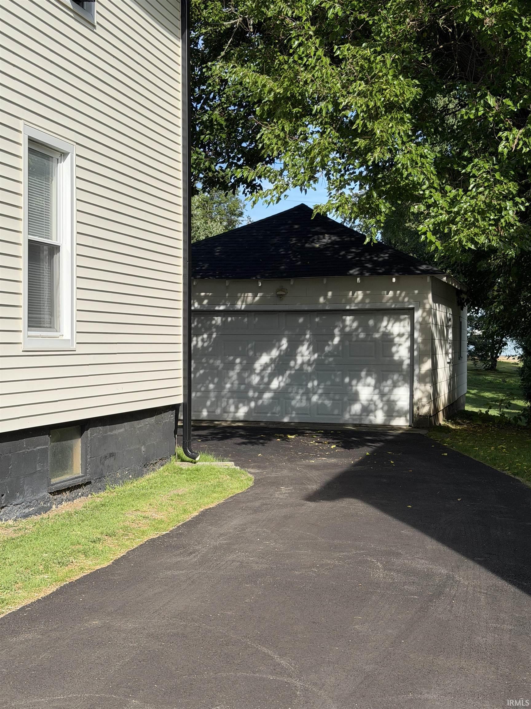 Detached two-car garage beside a light-sided house, with a broad asphalt driveway under leafy trees.