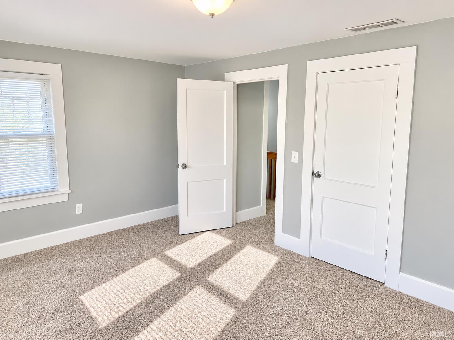 Empty bedroom with gray walls, beige carpet, and sunlight through a window with blinds.