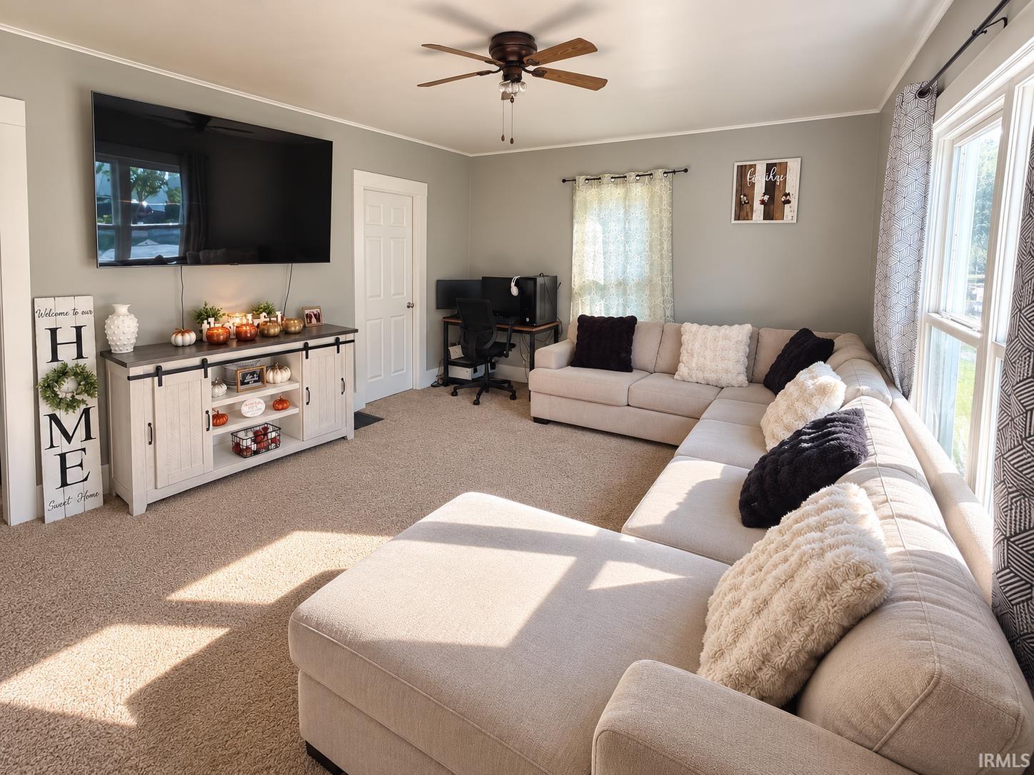 Furnished living room with a beige sectional, wall-mounted television, and sunlight streaming through tall windows.
