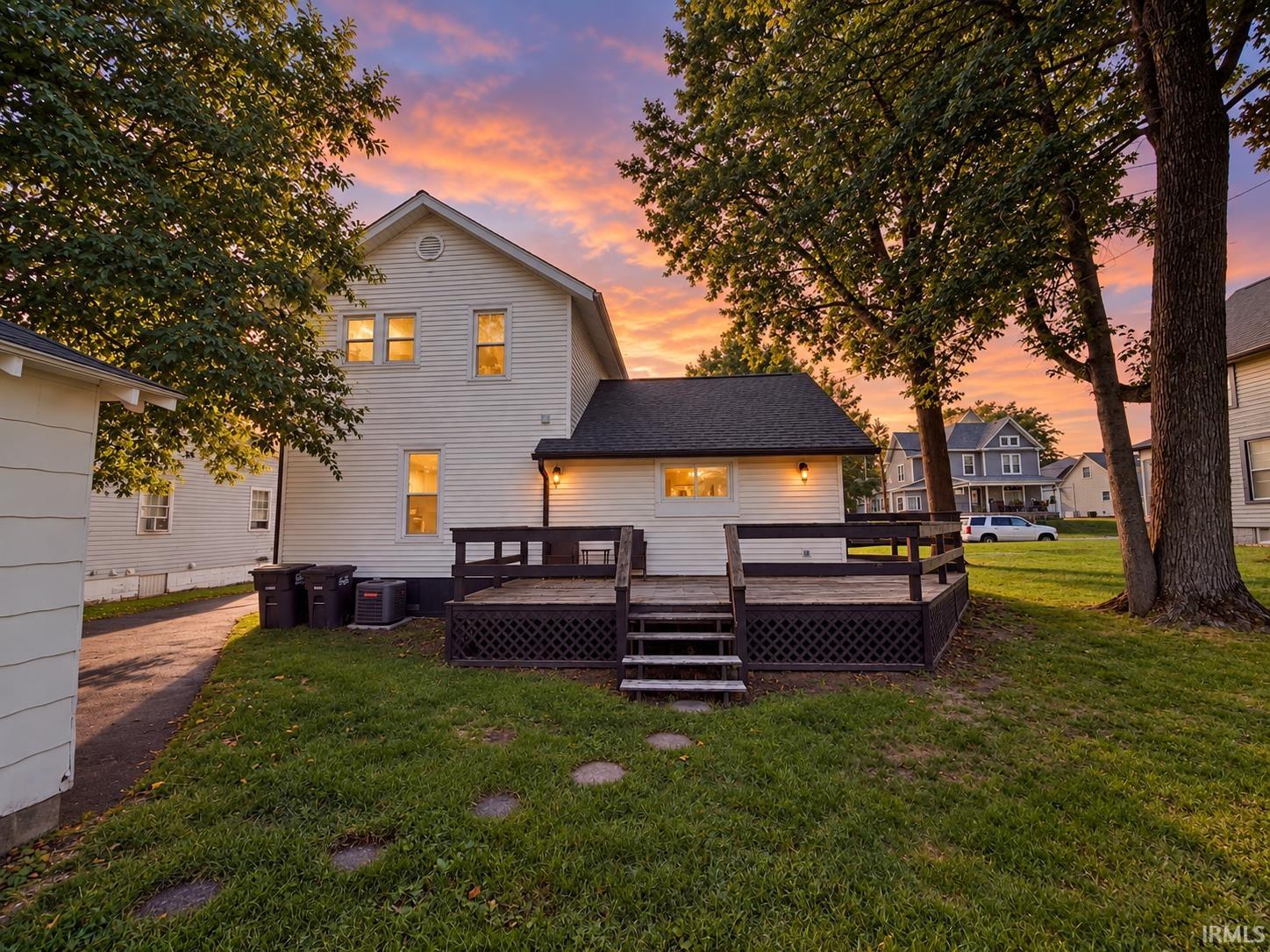 Rear of a light-colored two-story house with a raised deck and lawn beneath a vivid sunset.