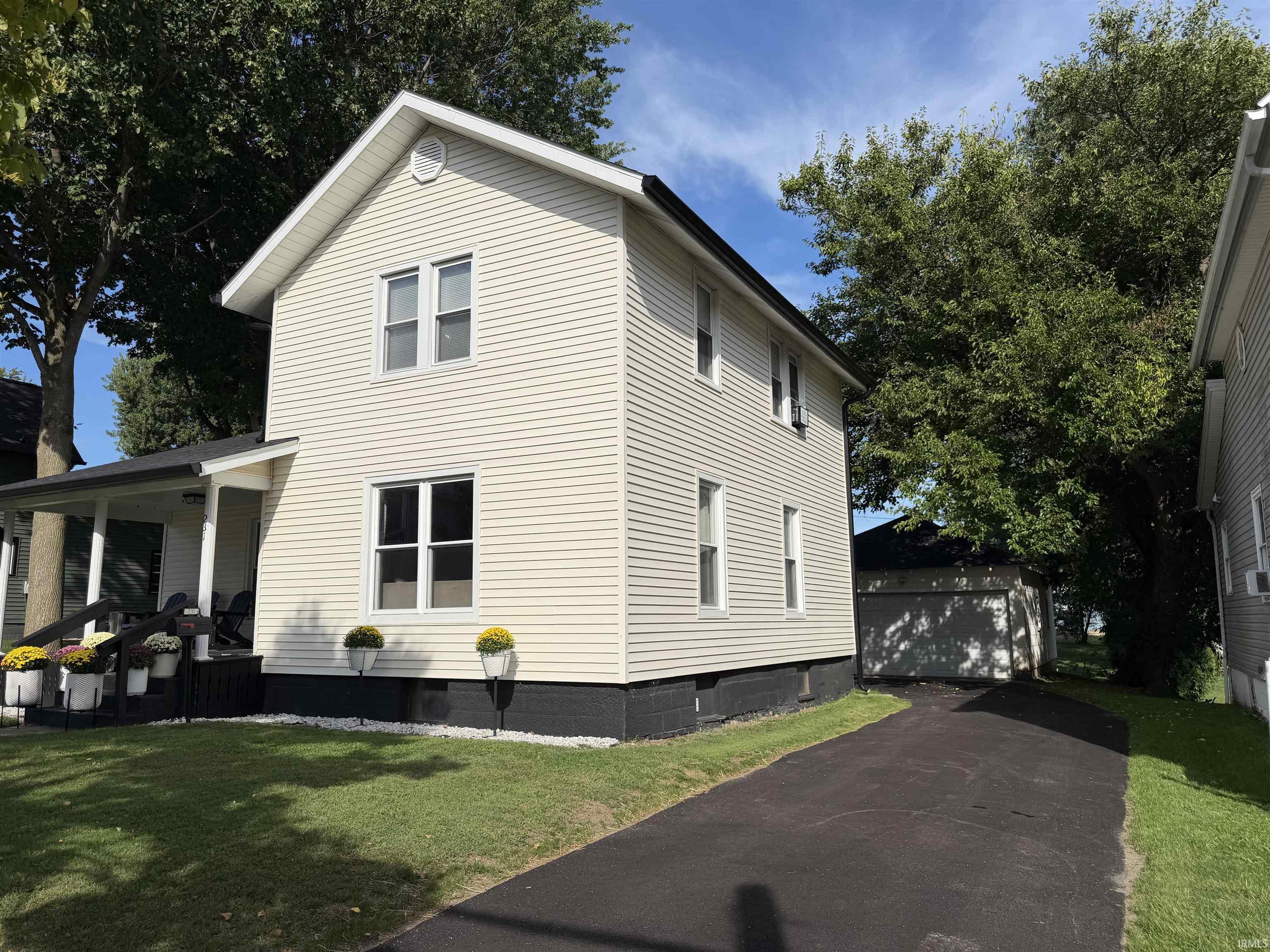 Cream two-story house with a covered porch, mature trees, and driveway leading to a detached garage.