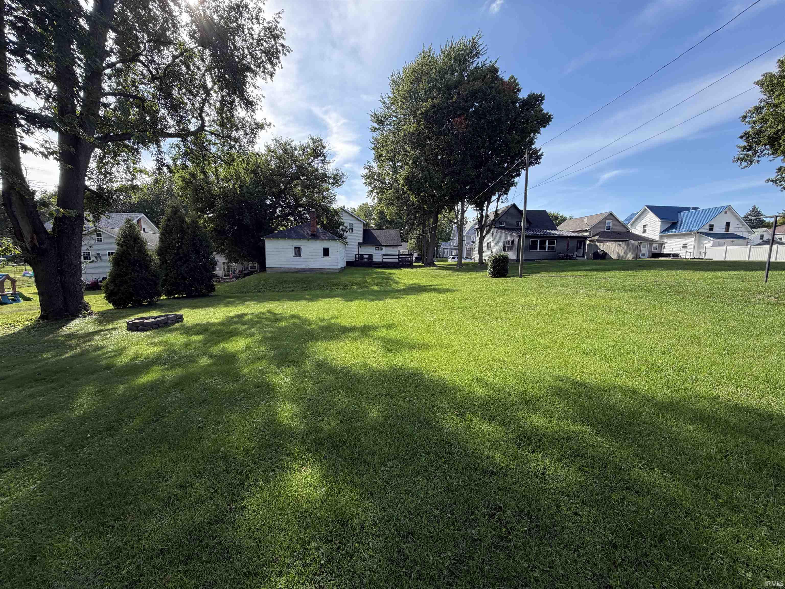 Spacious grassy backyard with mature trees, a stone fire pit, and neighboring homes beyond.