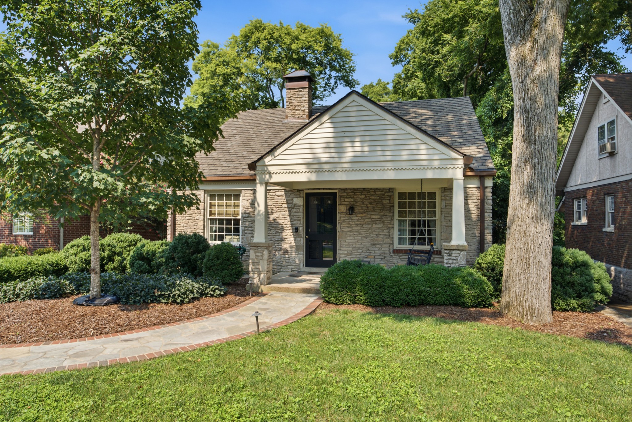 Stone house with a covered porch, mature trees, and a curved walkway through the front yard.