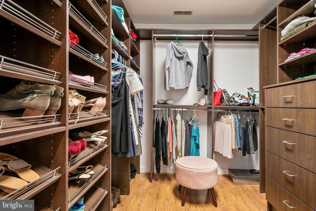 Walk-in closet with dark wood shelving, shoe racks, hanging clothes, and a round ottoman.