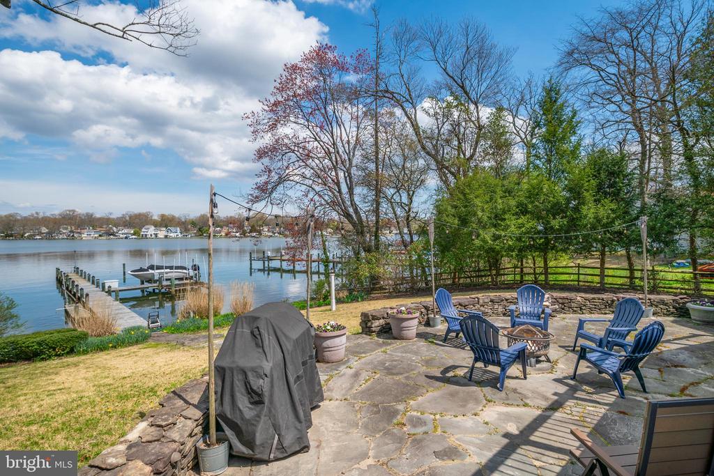 Stone waterfront patio with Adirondack chairs and a fire pit overlooking a dock and calm water.