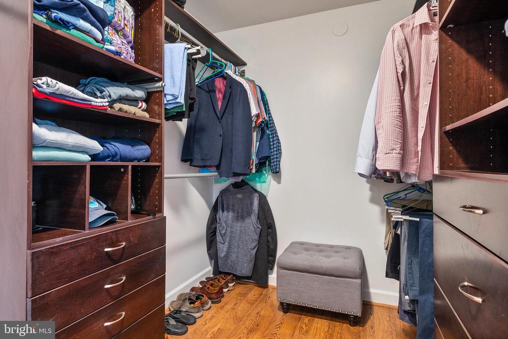 Walk-in closet with dark wood built-ins, hanging clothes, and a gray upholstered ottoman.