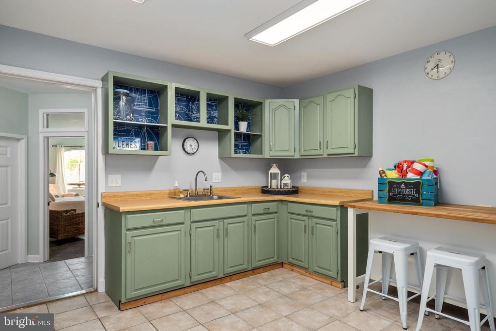 Sage-green kitchenette with wood countertops, open shelves, a sink, and two white stools.
