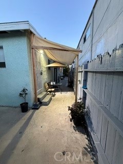 Narrow patio walkway with a fabric canopy, chairs, and potted plants beside a blue stucco building.