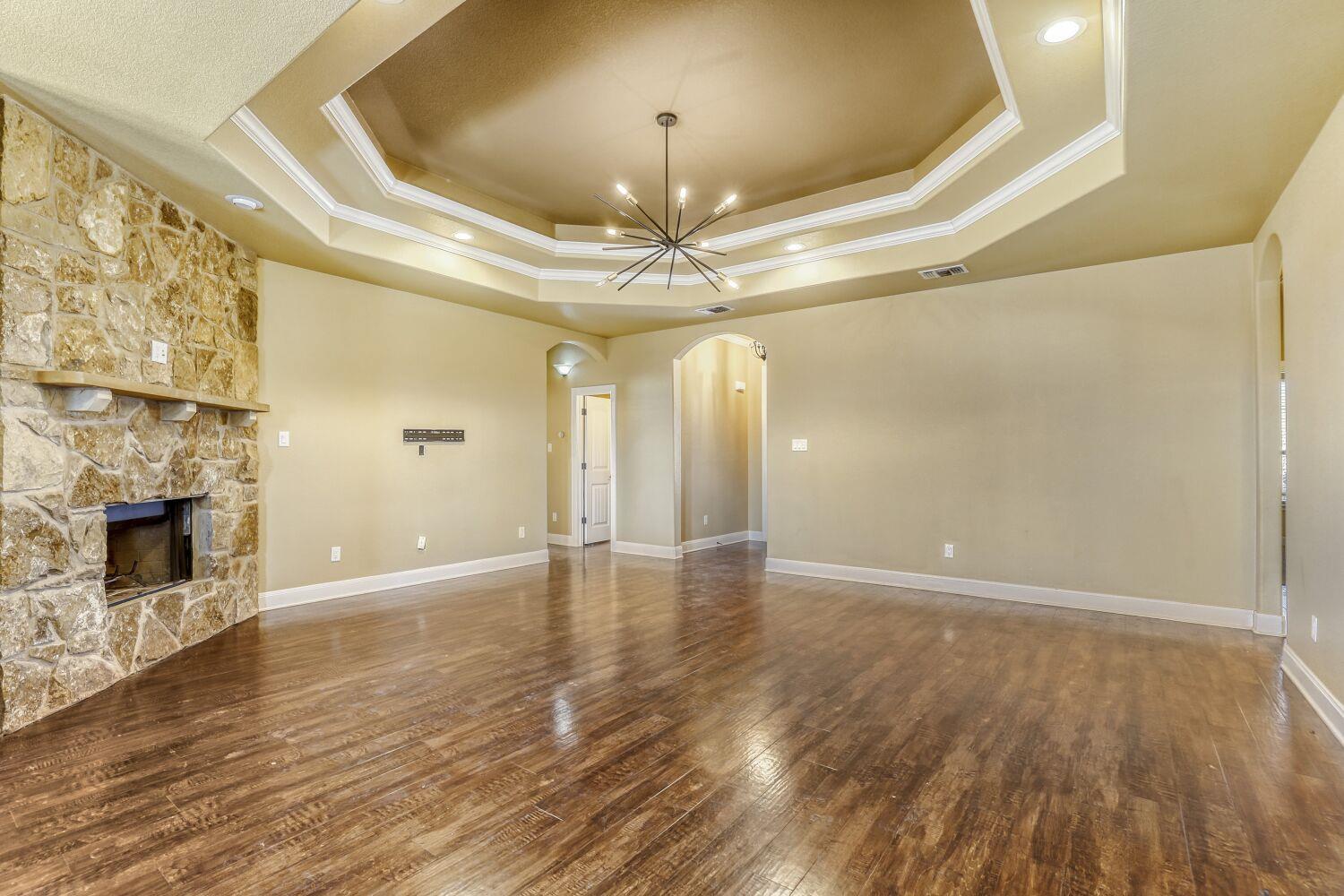 Empty living room with a stone fireplace, hardwood floors, and a tray ceiling with a modern chandelier.