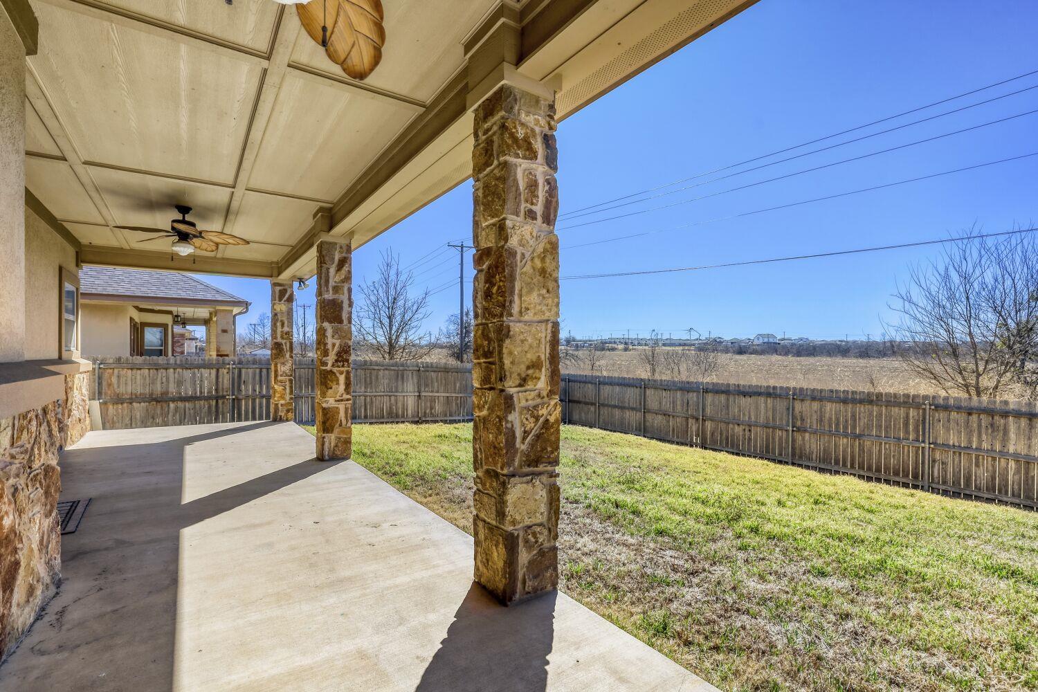 Covered patio with stone columns and ceiling fan overlooking a fenced backyard and open field.